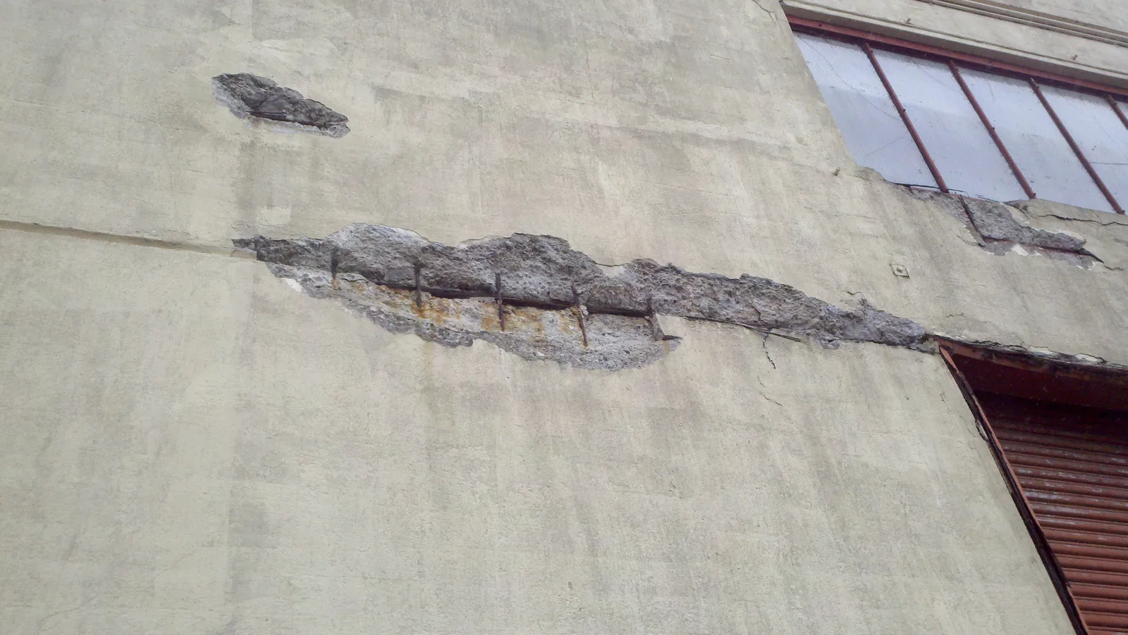 A long spalled strip in a concrete wall exposes a rust-coloured reinforcing line beneath the surface