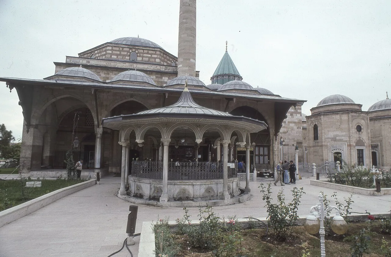 A roofed courtyard fountain in front of the domes and turquoise tower of the Mevlana Museum in Konya