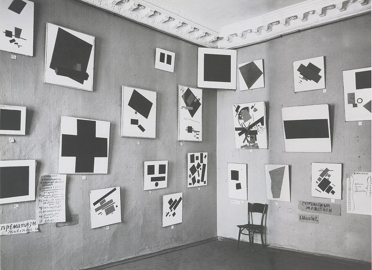 Black-and-white photograph of closely hung geometric paintings with Black Square high in a corner